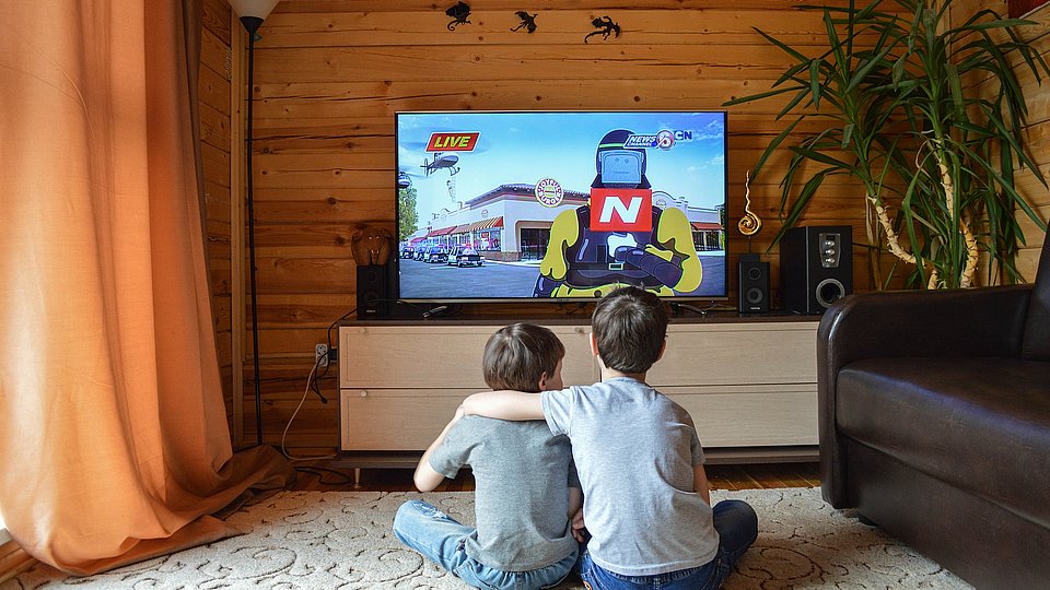 Zwei Jungen sitzen auf dem Teppich vor dem Fernseher, zu sehen ist ihre Rückenansicht. Der Ältere hat dem Jüngeren einen Arm um die Schulter gelegt, im Fernsehen läuft ein Action-Zeichentrickfilm. 