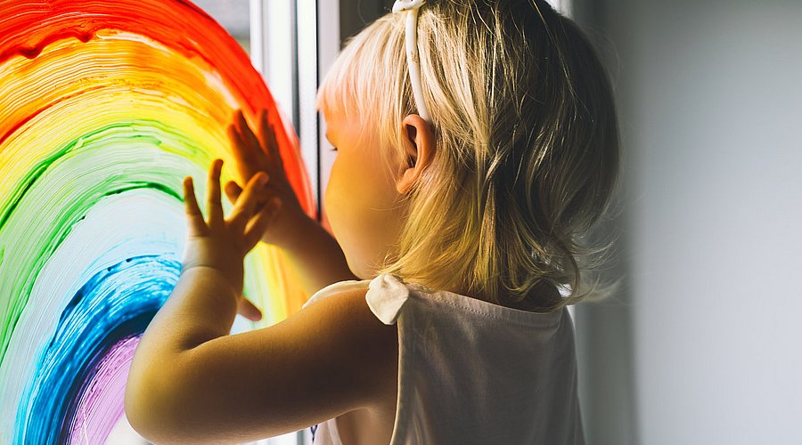Kita-Kind malt mit Fingerfarben einen Regenbogen ans Fenster. 