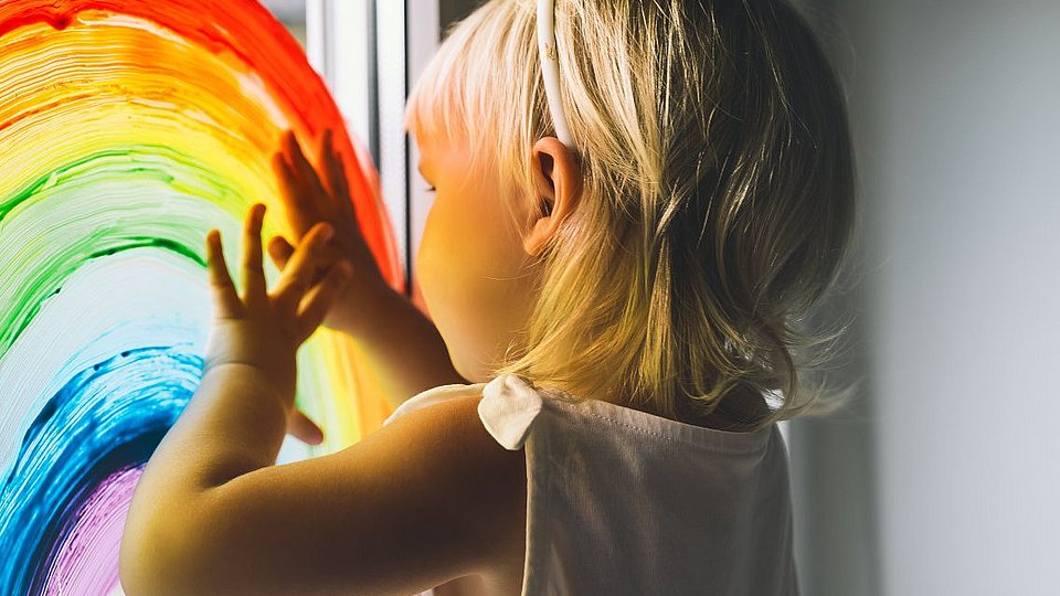 Kita-Kind malt mit Fingerfarben einen Regenbogen ans Fenster. 