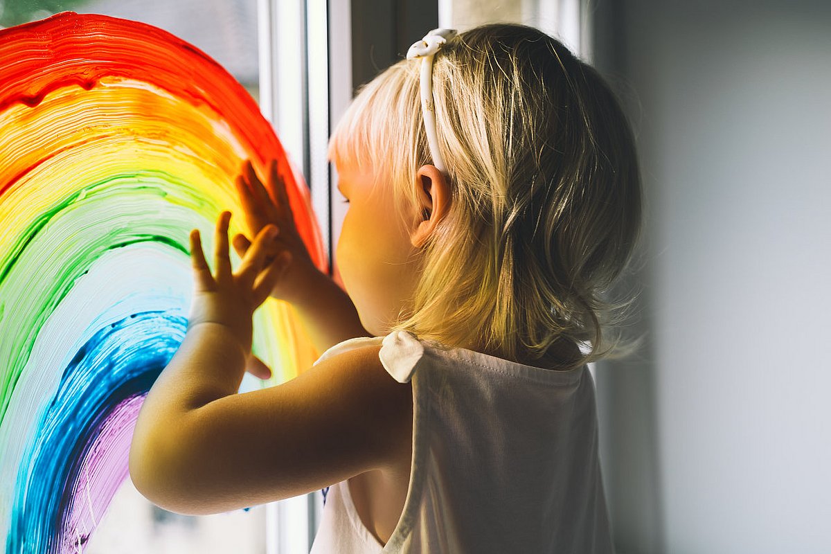Kita-Kind malt mit Fingerfarben einen Regenbogen ans Fenster. 