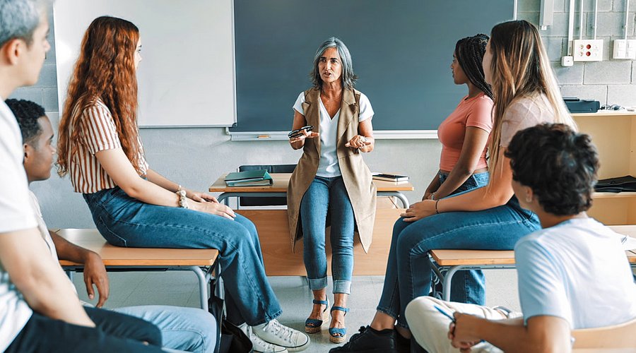 In einer Schulklasse steht eine Lehrerin am Pult und spricht mit Schüler*innen. 