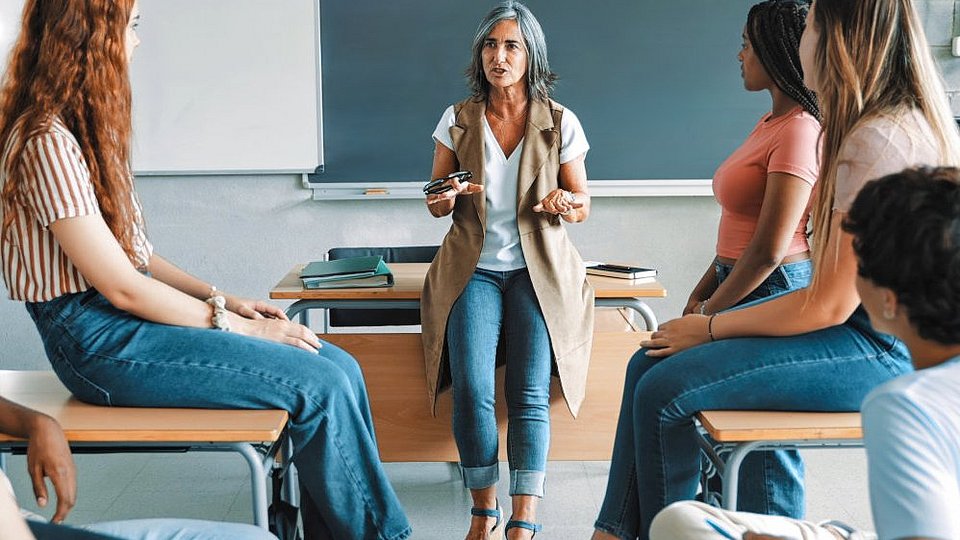 In einer Schulklasse steht eine Lehrerin am Pult und spricht mit Schüler*innen. 