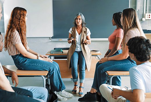 In einer Schulklasse steht eine Lehrerin am Pult und spricht mit Schüler*innen. 