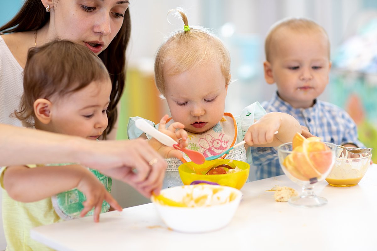 Erzieherin sitzt mit drei Kleinkindern am Tisch und hilft ihnen beim Essen.