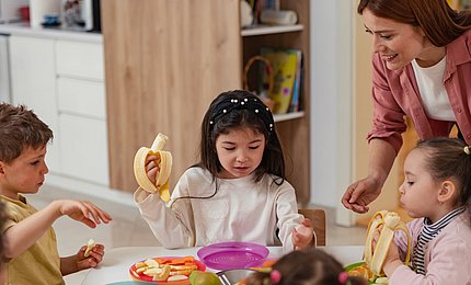 Eine Gruppe Kita-Kinder sitzt gemeinsam am Tisch und isst Obst, während eine pädagogische Fachkraft sie begleitet und anleitet. 