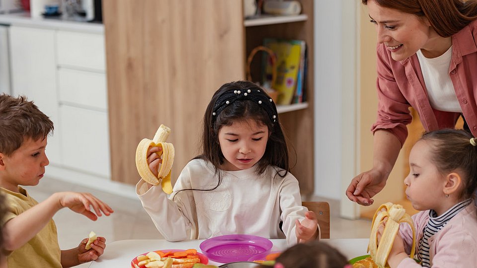 Eine Gruppe Kita-Kinder sitzt gemeinsam am Tisch und isst Obst, während eine pädagogische Fachkraft sie begleitet und anleitet. 