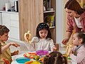 Eine Gruppe Kita-Kinder sitzt gemeinsam am Tisch und isst Obst, während eine pädagogische Fachkraft sie begleitet und anleitet. 
