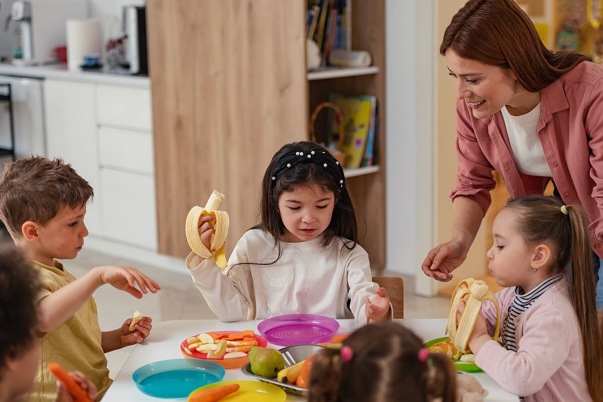 Eine Gruppe Kita-Kinder sitzt gemeinsam am Tisch und isst Obst, während eine pädagogische Fachkraft sie begleitet und anleitet. 