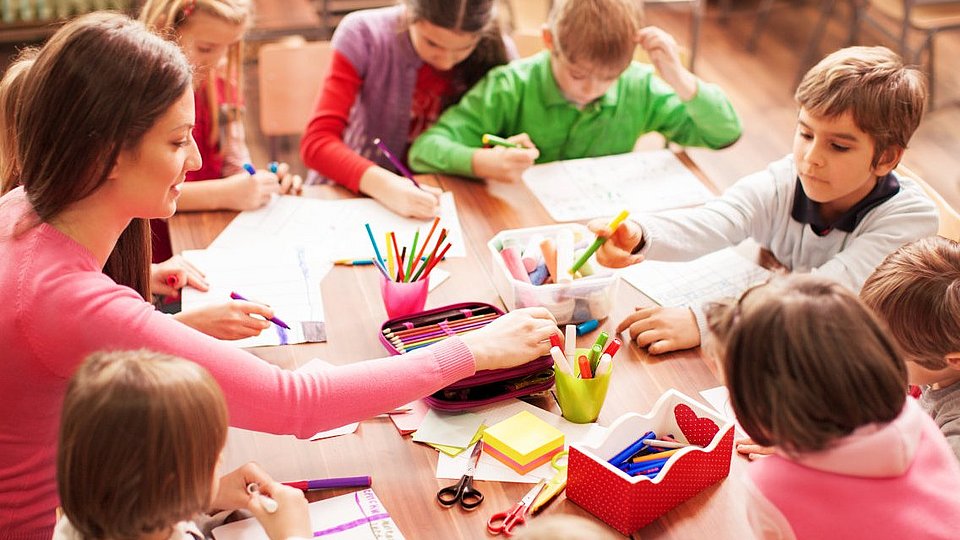 In einem Klassenraum sitzen Schüler*innen mit ihrer Lehrerin an einem Tisch und malen und basteln. 