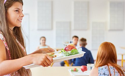 Schulmädchen steht mit einem Tablett in der Mensa. 