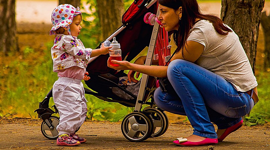 Ein bunt gekleidetes Kleinkind mit Sonnenhütchen steht neben seinem Kinderwagen, vor ihm kniet seine sommerlich gekleidete Mutter. Sie reicht dem Kind eine Babyflasche mit einem Getränk. 