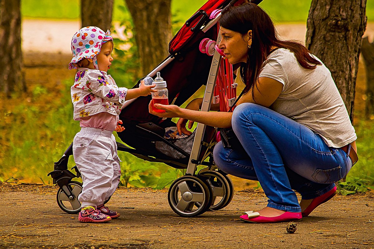 Ein bunt gekleidetes Kleinkind mit Sonnenhütchen steht neben seinem Kinderwagen, vor ihm kniet seine sommerlich gekleidete Mutter. Sie reicht dem Kind eine Babyflasche mit einem Getränk. 