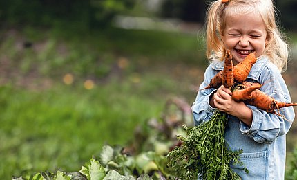 Ein kleines Mädchen hält einen Bund Möhren in der Hand und lächelt verschmitzt.
