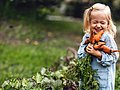 Ein kleines Mädchen hält einen Bund Möhren in der Hand und lächelt verschmitzt.