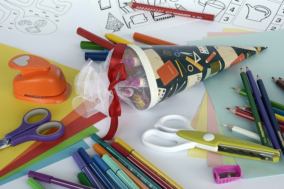 Auf einem Tisch liegen Scheren, Papier, Buntstifte und eine kleine Schultüte. Alles ist für Erstklässler*innen geeignet.