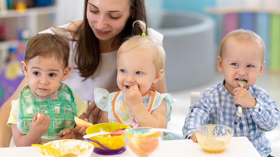 Kindertagespflegeperson sitzt mit Kleinkindern am Tisch und hilft ihnen beim Essen.