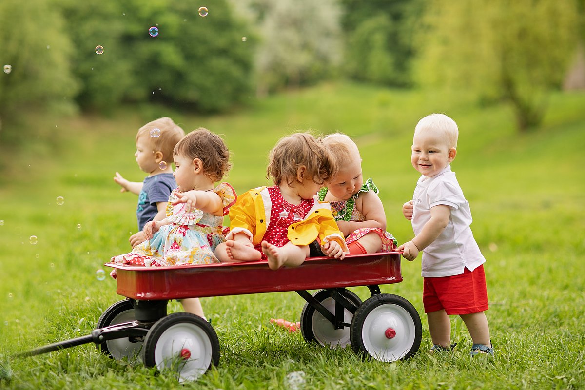 Eine Gruppe von fünf Kleinkindern sitzt und steht um einen roten Bollerwagen auf einer Wiese herum.