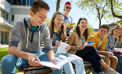 Jugendliche Schüler*innen sitzen mit ihren Schulunterlagen gemeinsam auf einer Bank. 