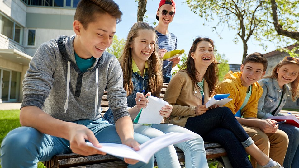 Jugendliche Schüler*innen sitzen mit ihren Schulunterlagen gemeinsam auf einer Bank. 