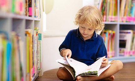 Kind liest vor einem Bücherregal in einem Buch.