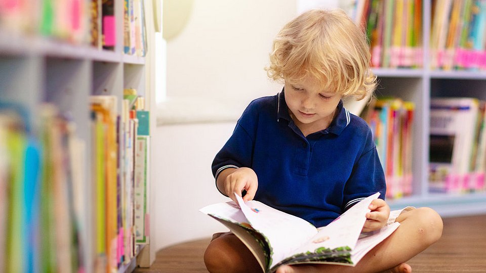 Kind liest vor einem Bücherregal in einem Buch.