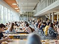 In einer Schulmensa sitzen viele Schüler*innen an Tischen und essen.