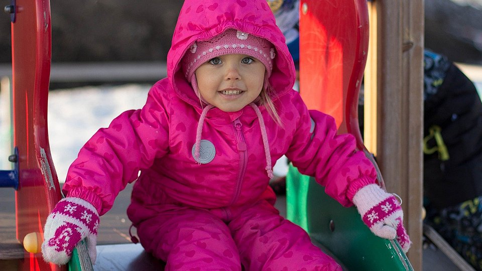 Ein kleines Mädchen im rosa Skianzug sitzt auf einer Rutsche auf einem verschneiten Spielplatz.