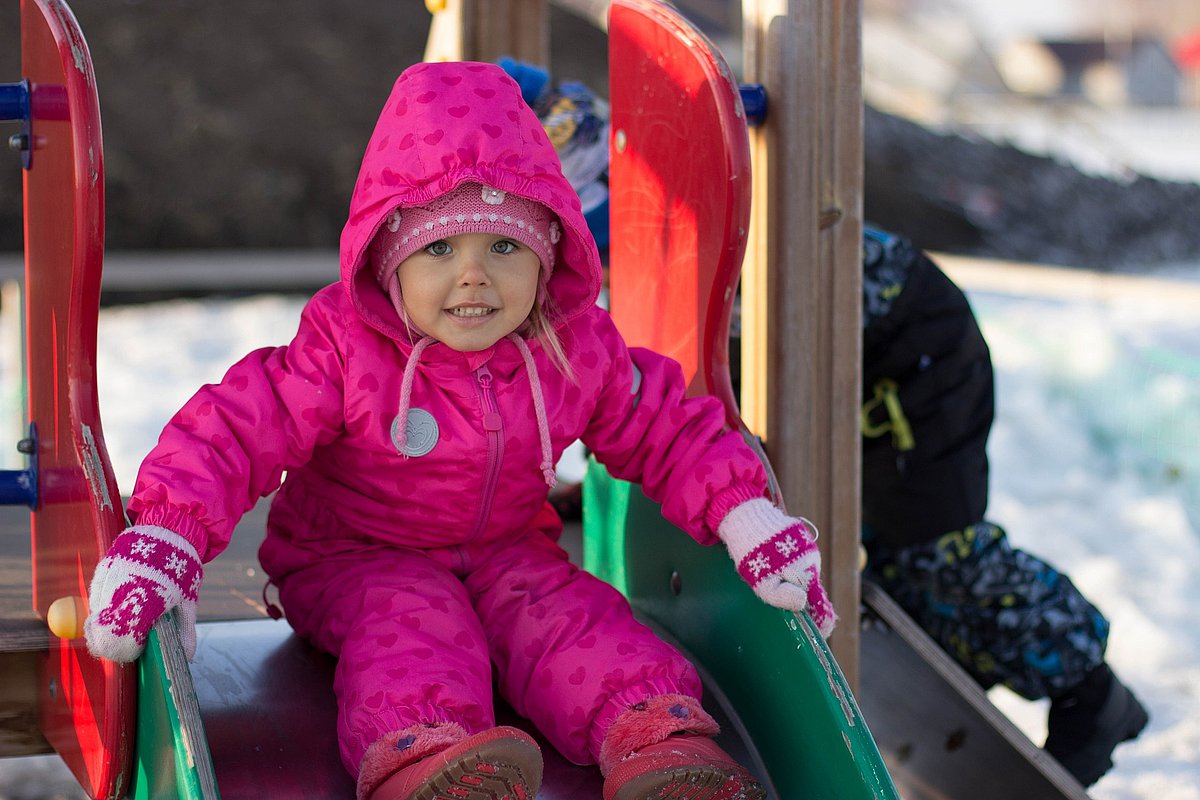 Ein kleines Mädchen im rosa Skianzug sitzt auf einer Rutsche auf einem verschneiten Spielplatz.