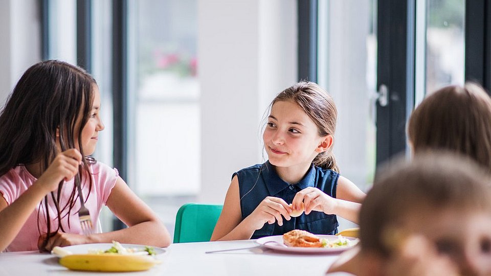 Zwei Mädchen im Grundschulalter am Tisch einer Mensa, das Mittagessen vor sich.