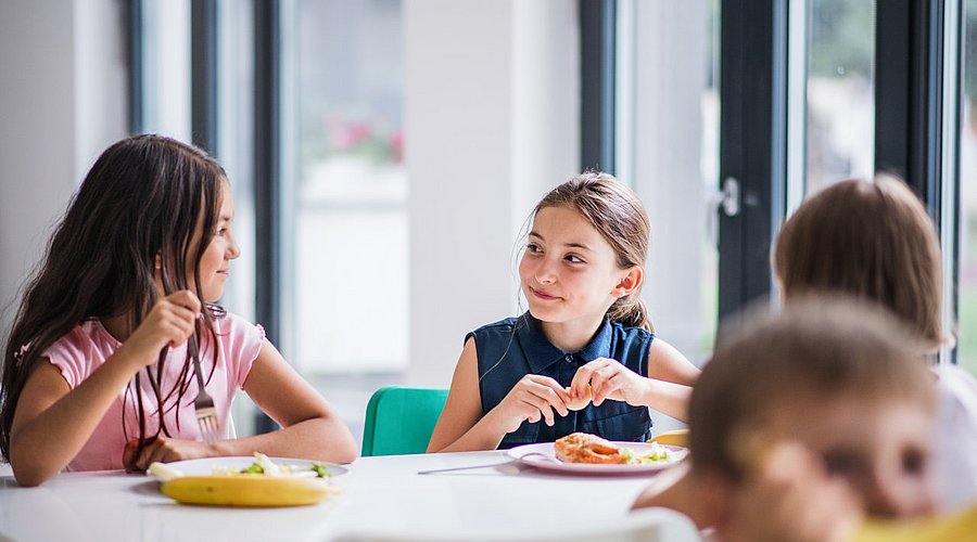 Zwei Mädchen im Grundschulalter am Tisch einer Mensa, das Mittagessen vor sich.