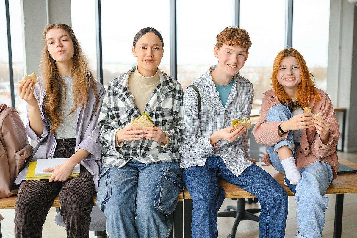 Teenager sitzen in der Schulmensa auf einem Tisch und snacken.