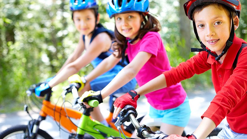 Drei Kinder mit Helm sitzen auf dem Fahrrad und gucken in die Kamera