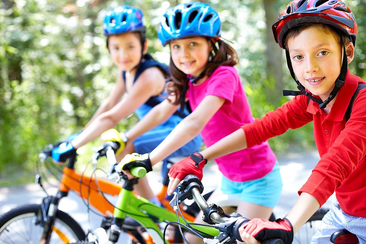 Drei Kinder mit Helm sitzen auf dem Fahrrad und gucken in die Kamera