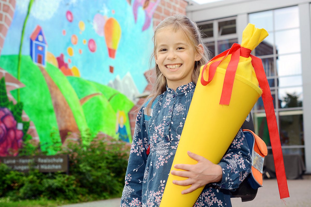 Ein Mädchen mit Schultüte steht lachend vor dem Schultor. 