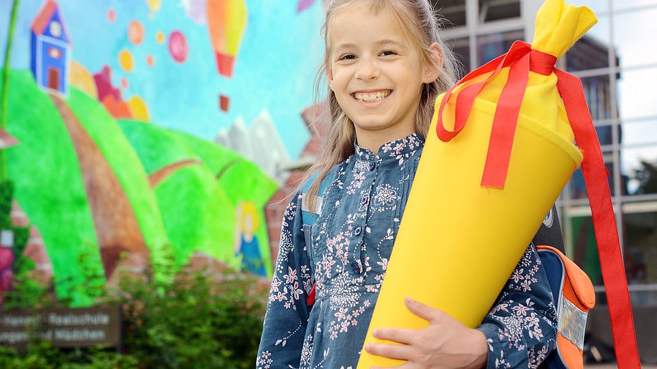 Ein Mädchen mit Schultüte steht lachend vor dem Schultor. 