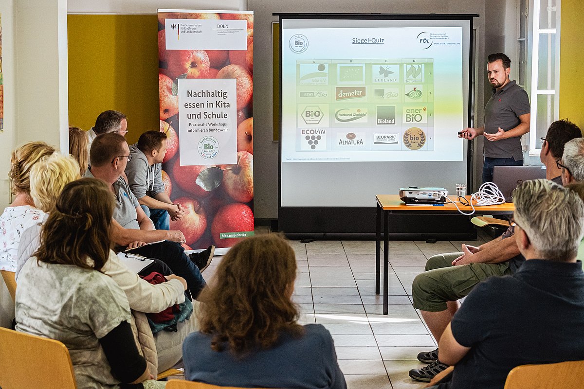 Workshop-Teilnehmende sitzen in einer Runde und hören dem Seminarleiter zu, der mitfhilfe einer Präsentation die Inhalte erklärt.
