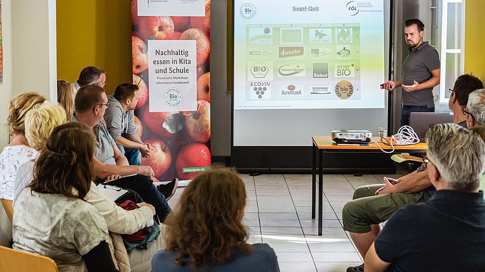 Workshop-Teilnehmende sitzen in einer Runde und hören dem Seminarleiter zu, der mitfhilfe einer Präsentation die Inhalte erklärt.