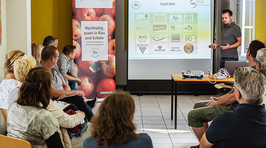 Workshop-Teilnehmende sitzen in einer Runde und hören dem Seminarleiter zu, der mitfhilfe einer Präsentation die Inhalte erklärt.