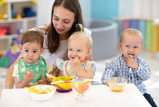 Drei Kleinkinder essen am Tisch mit Betreuerin