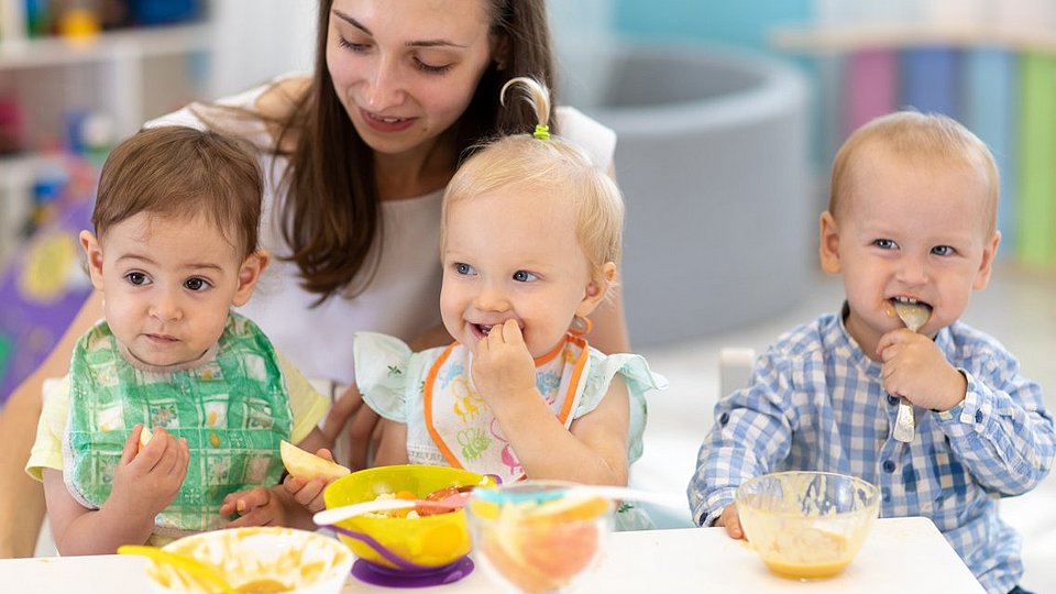 Kindertagespflegeperson sitzt mit Kleinkindern am Tisch und hilft ihnen beim Essen.