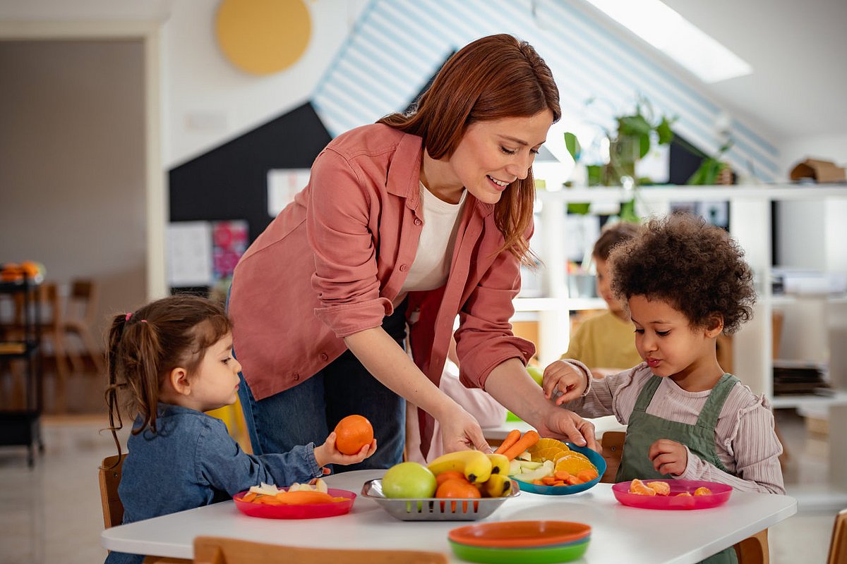 Kita-Kinder essen Obst, eine pädagogische Fachkraft reicht einen Teller an.