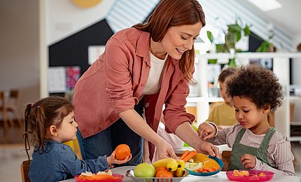 Kita-Kinder essen Obst, eine pädagogische Fachkraft reicht einen Teller an.