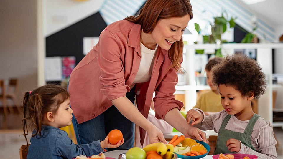 Kita-Kinder essen Obst, eine pädagogische Fachkraft reicht einen Teller an.