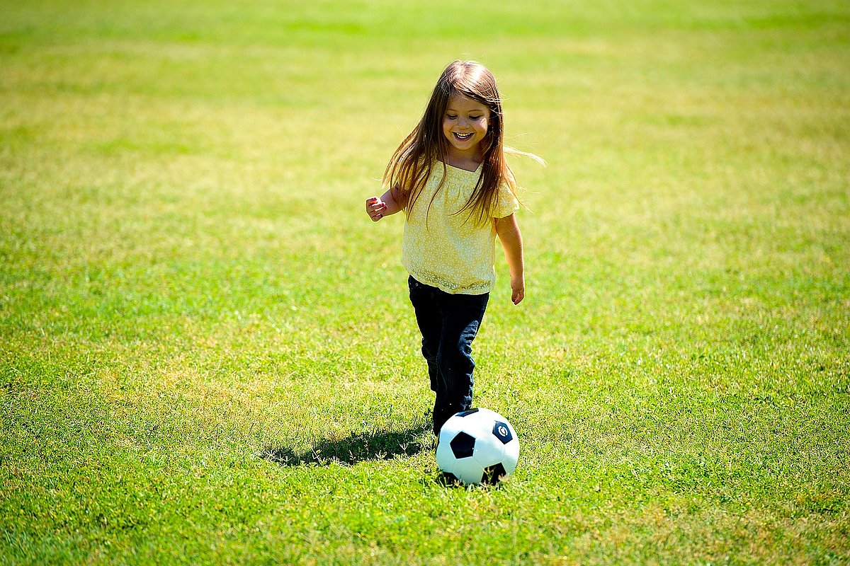 Kleines Mädchen spielt bei Sommerwetter auf dem Rasen mit einem Fußball.