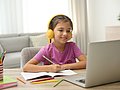 Ein Schulkind sitzt beim Home Schooling vor dem Laptop. Es hat gelbe Kopfhörer auf, vor ihm liegt ein geöffnetes Schulheft. 