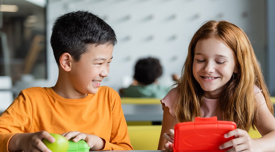 Zwei Schulkinder sitzen in der Schulklasse und essen ihr Frühstück aus der Brotdose. 