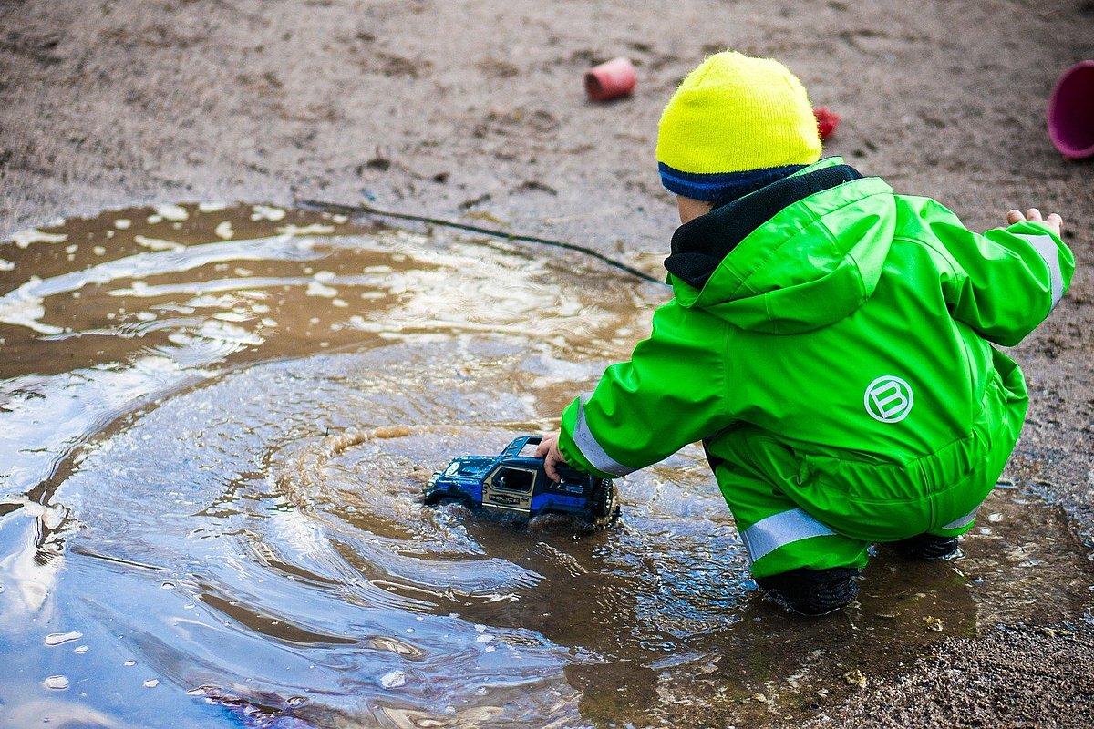 Ein Kind im Regenanzug spielt mit Auto in einer Pfütze.