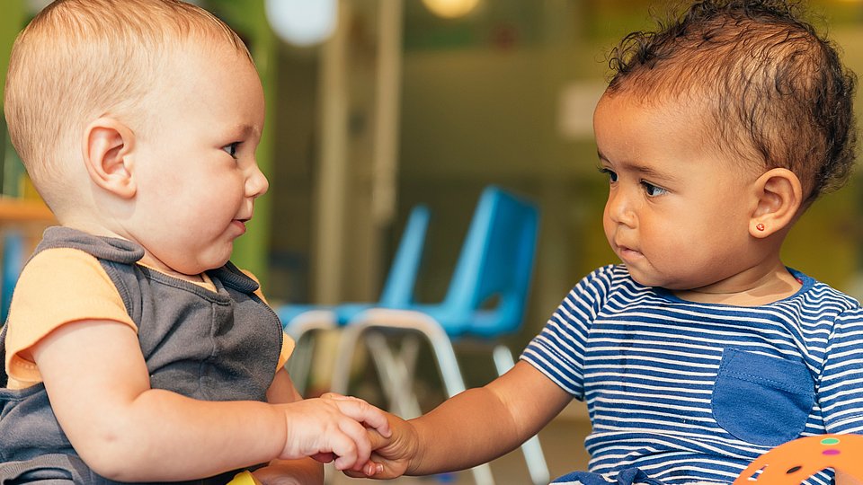Zwei Kleinkinder sitzen auf dem Boden, schauen sich an und geben sich gegenseitig ein Spielzeug.