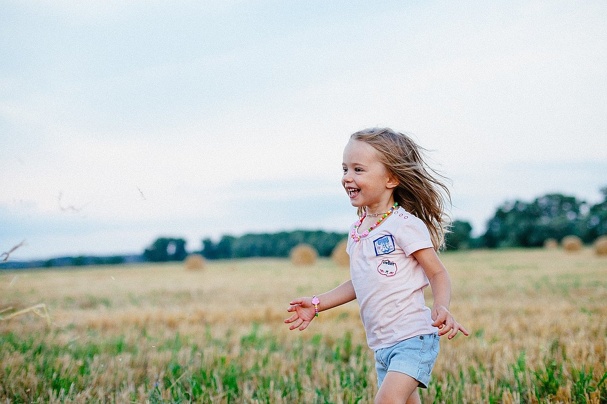 Kleines Mädchen mit langen blonden Haaren in T-Shirt und kurzer Jeanshose läuft am Rande eines sommerlichen Getreidefeldes.