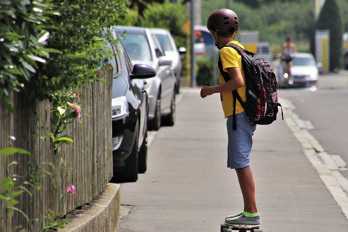 Ein Junge fährt auf einem Skateboard über den Bürgersteig. Er trägt kurze Hosen und Helm.